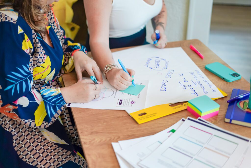 Renia Carsillo and Stephanie Veraghen collaborating at a strategic planning retreat, writing ideas on a large paper with colorful pens and sticky notes